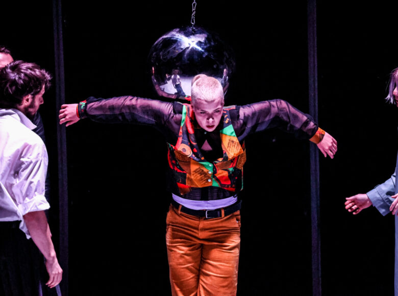 Zdjęcie. Grupa osób w kolorowych strojach stoi na ciemnej scenie. Za nimi, na łańcuchu wisi szklana kula.