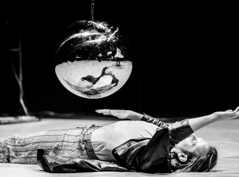 Czarno-białe zdjęcie. Mężczyzna leży na podłodze. Jedną rękę unosi nad siebie, druga kieruje wzdłuż tułowia. Nad nim przelatuje zawieszona na łańcuchu skalana kula.
