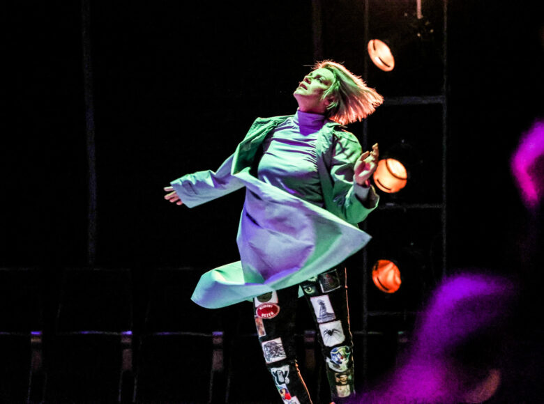 Zdjecie. Kobieta patrzy w górę i skacze. Ma rozwiane, krótkie blond włosy. Jest ubrana w jasną koszulę i płaszcz w takim samym odcieniu oraz czarne spodnie oklejone różnymi grafikami, przedstawiającymi ikony popkultury. Scena jest ciemna, świecą się trzy pomarańczowe reflektory.