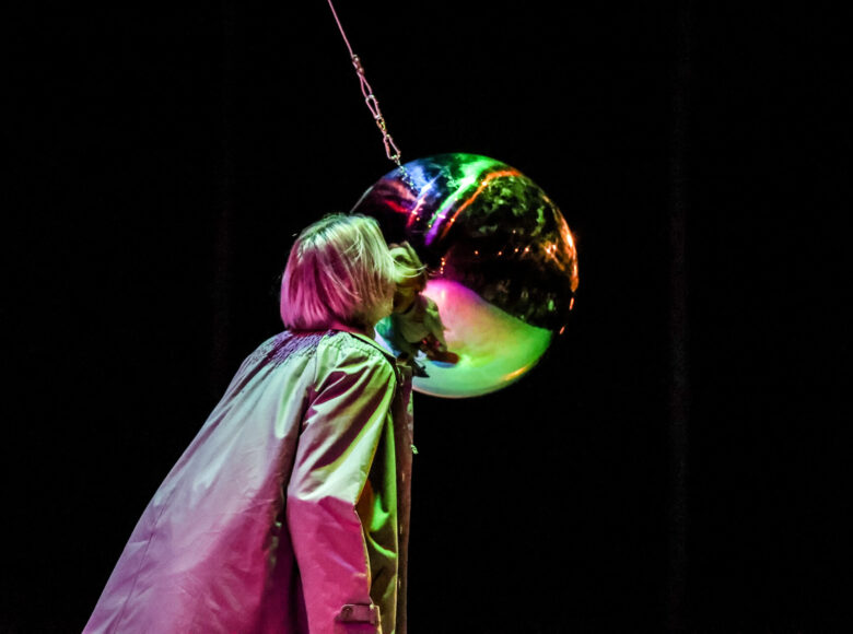 Zdjęcie. Ciemna scena. Kobieta stoi tyłem. Zbliża twarz do szklanej kuli zawieszonej na łańcuchu.
