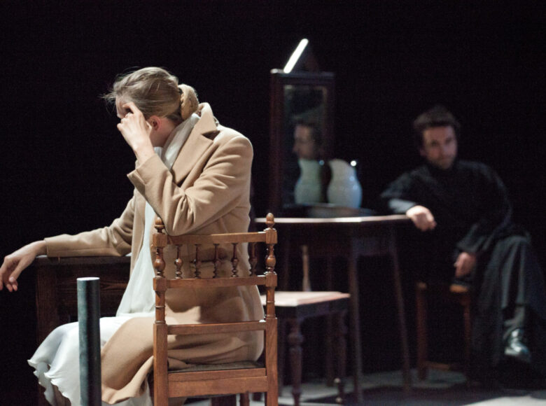 Zdjęcie. Scena ze spektaklu. Kobieta w białej sukience i beżowym płaszczu siedzi bokiem na drewnianym krześle. Za nią niewyraźna sylwetka mężczyzny w sutannie, który siedzi przy stoliku.