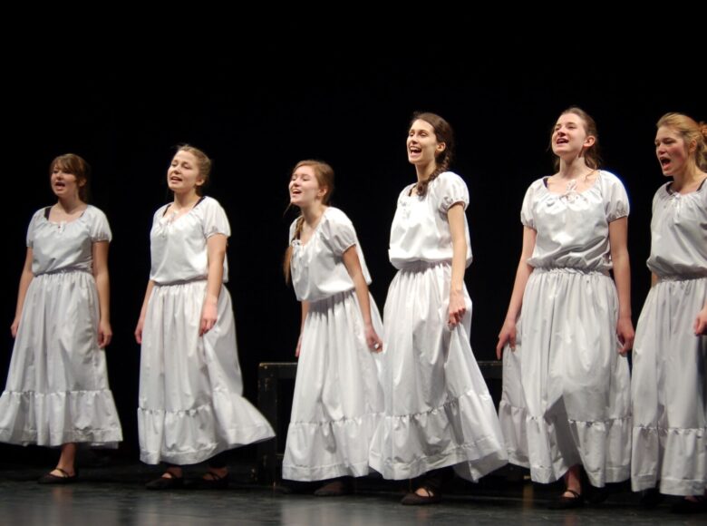 Zdjęcie ze spektaklu. 6 dziewcząt. Mają w większości długie włosy splecione w warkocze lub związane w kucyk. Śpiewają. Są ubrane w białe sukienki.