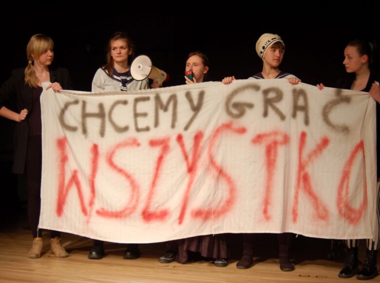 Zdjęcie ze spektaklu. Grupa pięciu stojących dziewcząt. Trzymają przed sobą prześcieradło z napisem „Chcemy grać wszystko”. Dwie są ubrane w stroje marynarskie. Trzy pozostałe są ubrane na czarno. dziewczyna na środku trzyma megafon.