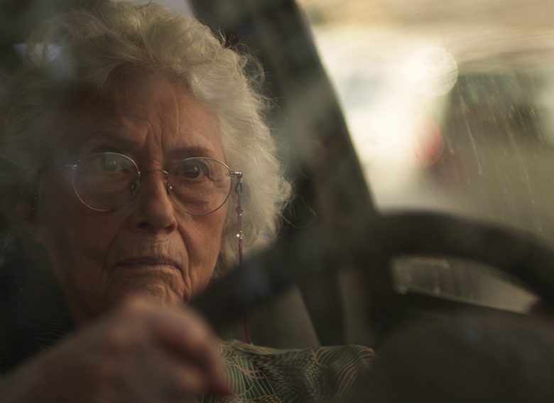 Kadr z filmu. Starsza, siwowłosa kobieta w okularach siedzi w aucie, obie ręce trzyma na kierownicy, patrzy z determinacją przed siebie. Ma zacięte usta.