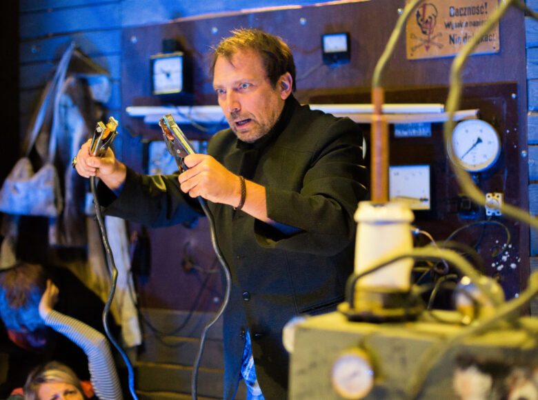 Zdjęcie. Scena ze spektaklu. Mężczyzna trzyma w rękach przewody rozruchowe. Jest przejęty. Za nim znajduje się elektroniczna aparatura oraz tabliczka z napisem ostrożność, nie dotykać.