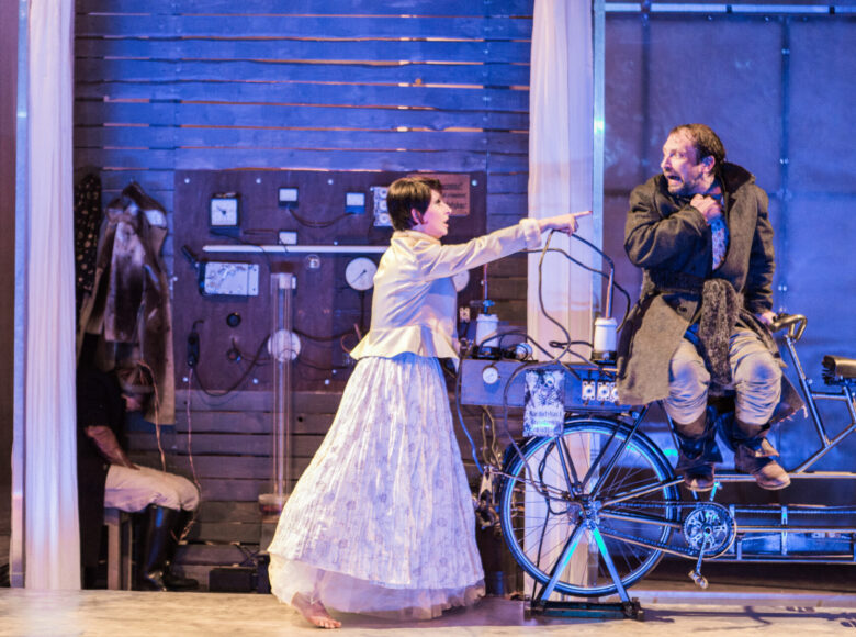 Zdjęcie. Scena ze spektaklu. Mężczyzna siedzi na opartym na stojaku rowerze. Patrzy w bok, na kobietę, w stroju z epoki, która wskazuje na niego palcem. Jest przerażony.