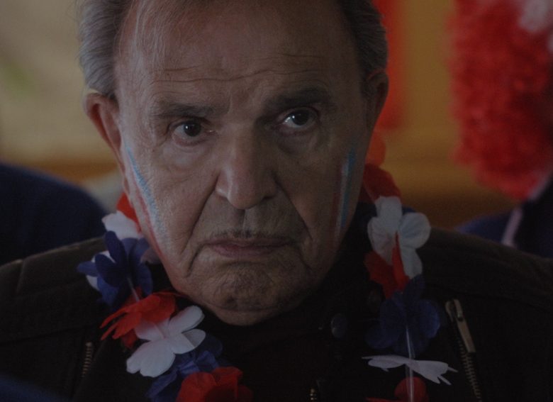Kadr z filmu. Starszy mężczyzna z czerwono-biało-niebieskimi kwiatami na szyi i namalowanymi barwami na policzkach, siedzi wśród innych osób.