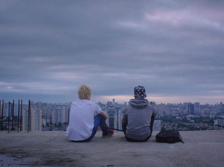 Zdjęcie. Kadr z filmu. Dwóch młodych chłopaków siedzi na betonowej kładce. Jeden z nich ma na sobie biały podkoszulek, a drugi szarą bluzę i granatowa czapkę z daszkiem, obok niego leży czarny plecak. Są odwróceni tyłem. Przed nimi rozciąga się widok na miasto pełne wieżowców. Niebo jest zachmurzone.