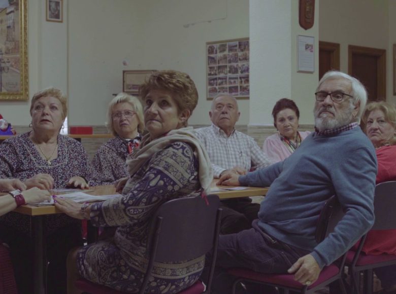 Kadr z filmu. Grupa kilkunastu starszych osób, siedzi przy długim stole w sali. Wszyscy z zainteresowaniem patrzą na coś lub kogoś poza kadrem.