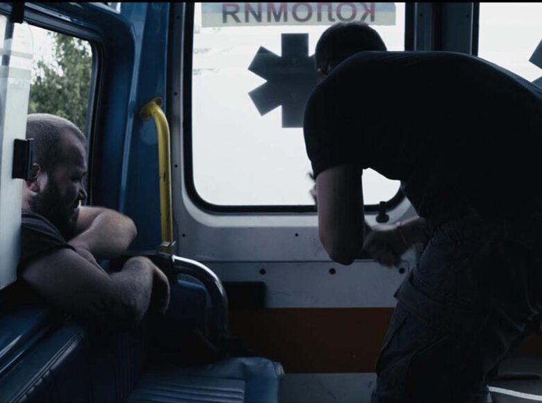 Zdjęcie. Kadr z filmu. Wnętrze karetki pogotowia, W środku znajduje się odwrócony tyłem mężczyzna. Przez okno od zewnątrz wychyla się drugi mężczyzna.