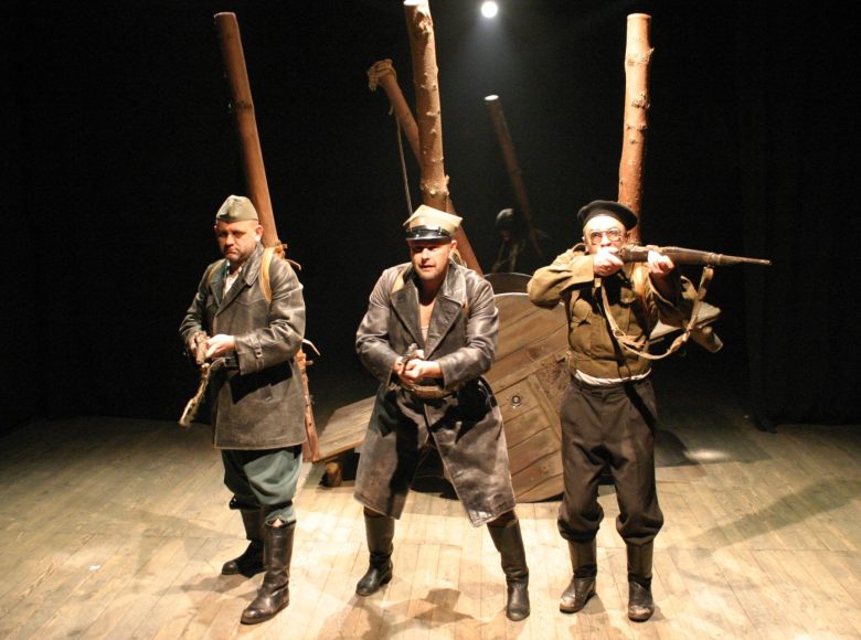 Zdjęcie. Scena ze spektaklu. Trzej mężczyźni w wojskowych strojach mierzą z karabinów. Za nimi znajduje się drewniany wóz i pnie drzew.