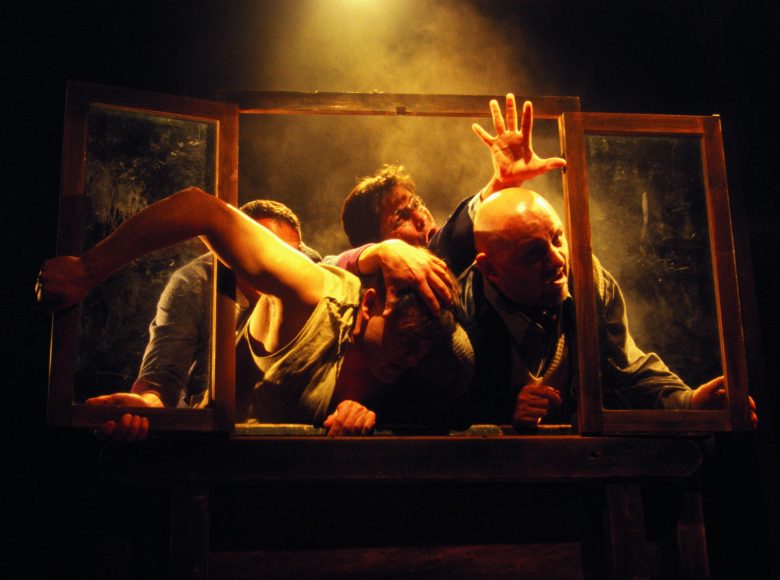 Zdjęcie. Scena ze spektaklu. Grupa mężczyzn wychyla się z drewnianej ramy okna. Przepychają się, mają przerażone miny, próbują uciekać. Za nimi dym. Na scenę pada pomarańczowe światło.