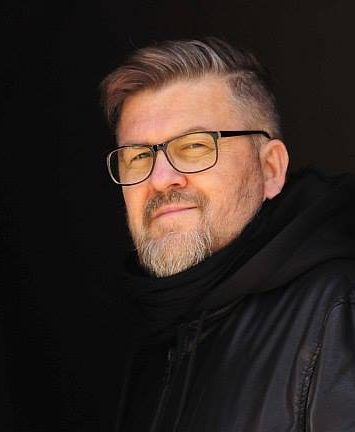 Zdjęcie. Portret mężczyzny w średnim wieku. Ma na sobie czarną kurtkę i bluzę z kapturem. Nosi okulary. Ma siwe, wystylizowane włosy i krótką bródkę. Lekko się uśmiecha. Tło czarne, jednolite.