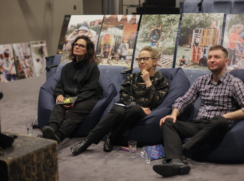 Zdjęcie. Dwie kobiety i mężczyzna siedzą na dużych, granatowych poduszkach. Patrza w jednym kierunku. Za nimi rozłożone są zdjęcia przedstawiające zajęcia w Rezerwacie Dzikich Dzieci. Na podłodze lezą szklanki i zeszyty.