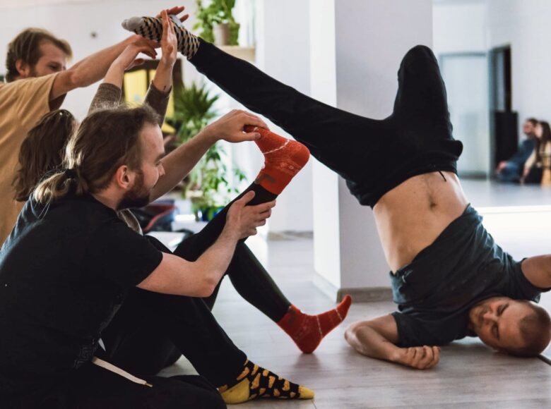 Zdjęcie. Grupa młodych mężczyzn, siedzą na ziemi, noszą kolorowe skarpetki. Jeden z nich stoi na głowie.