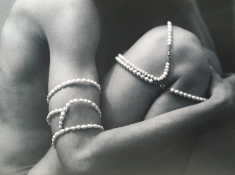 Czarno-białe zdjęcie. na zbliżeniu tułów nagiej kobiety w pozycji embrionalnej. Jej ramię i kolano oplatają sznury pereł.
