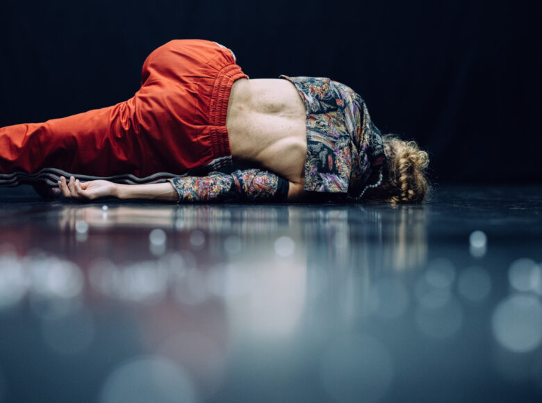Zdjęcie. Kobieta ubrana w pomarańczowe dresowe spodnie i krótką bluzkę w kolorowe wzory leży na scenie odwrócona tyłem. Ma odkrytą połowę pleców.