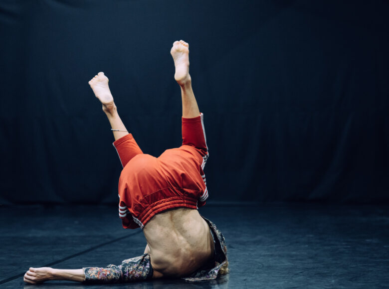 Zdjęcie. Zdjęcie. Kobieta ubrana w pomarańczowe dresowe spodnie i krótką bluzkę w kolorowe wzory znajduje się na scenie. Stoi na głowie.