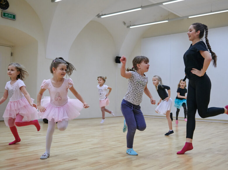Kolorowa fotografia. Wnętrze sali prób, w której w kolorowych spódniczkach i strojach tańczą kilkuletnie dziewczynki. Zajęcia prowadzi młoda kobieta.