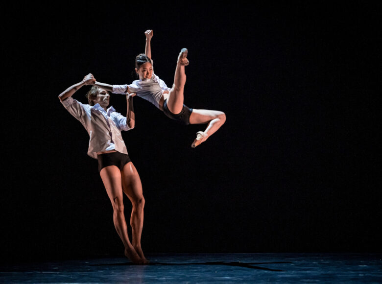 Kolorowa fotografia. Tańcząca para. Mężczyzna stoi wyprostowany, odchyla się lekko do tyłu. Rękami podtrzymuje kobietę, która wykonuje wysoki wyskok do góry.