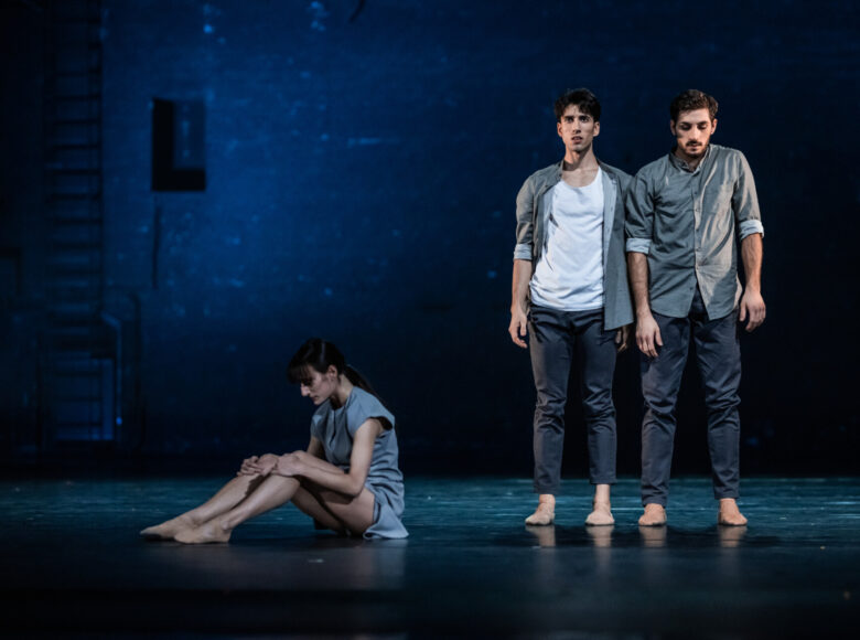 Kolorowa fotografia. Dwóch mężczyzn. Stoją ramie w ramie. Ubrani są w szare spodnie i szare koszule, maja bose stopy. Obok nich, na podłodze siedzi kobieta. Siedzi bokiem opierając ramiona o zgięte nogi. Patrzy w ziemie.