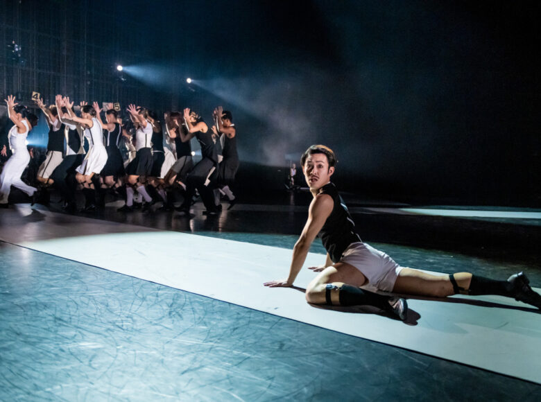 Kolorowa fotografia. Na scenie leży mężczyzna, podpiera się na ramionach i przez lewe ramię patrzy w stronę fotografa. W tle tańcząca grupa, światła reflektorów.