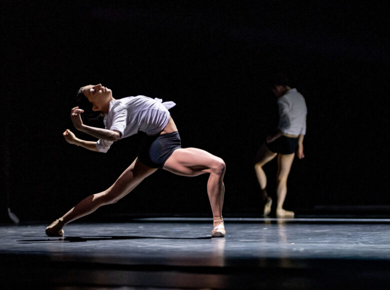 Kolorowa fotografia. Tańcząca kobieta. Stoi w rozkroku, plecy i ręce odchyla do tyłu. Światło reflektorów podkreśla jej umięśnione nogi. W tle rozmyta postać.