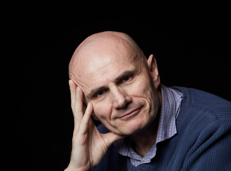 Zdjęcie. portret mężczyzny. Jest ubrany w jasną koszulę w kratkę i niebieski sweter. Na jednej ręce podpiera głowę. Mężczyzna jest łysy. Uśmiecha się. Tło czarne, jednolite.