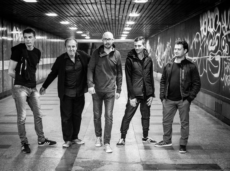 Czarno-białe zdjęcie. Pięciu mężczyzn znajduje się w podziemnym korytarzu. Na ścianach graffiti.