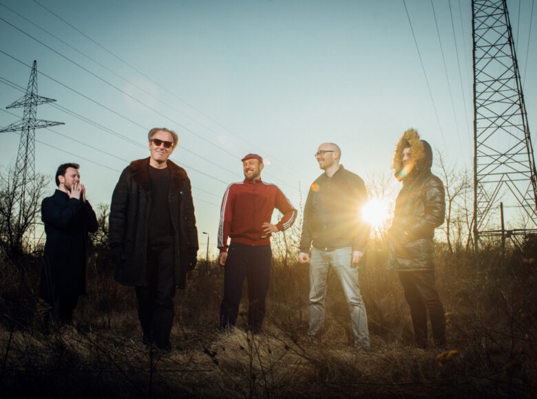Pięciu mężczyzn stoi pośrodku wczesnowiosennego pola. W tle przez sylwetki mężczyzn przebija się słońce, linie wysokiego napięcia oraz błękitne niebo.