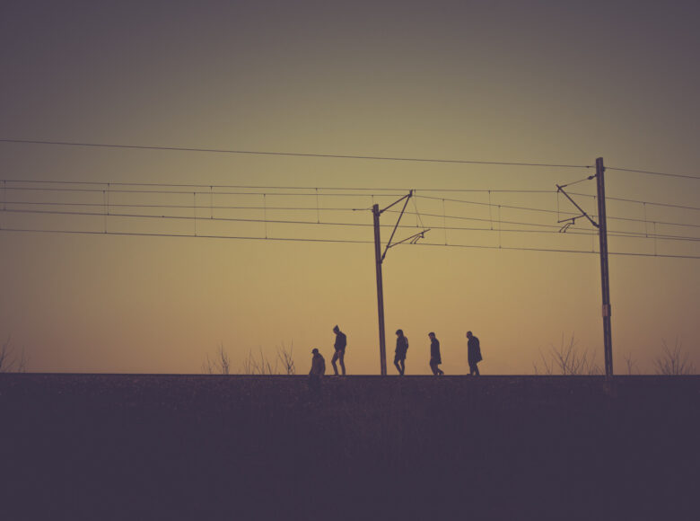 Na zdjęciu pięciu mężczyzn idzie horyzontalnie wzdłuż torów. Nad nim wzdłuż torów rozpościera się linia trakcji pociągów Jest schyłek dnia, słońce zachodzi.