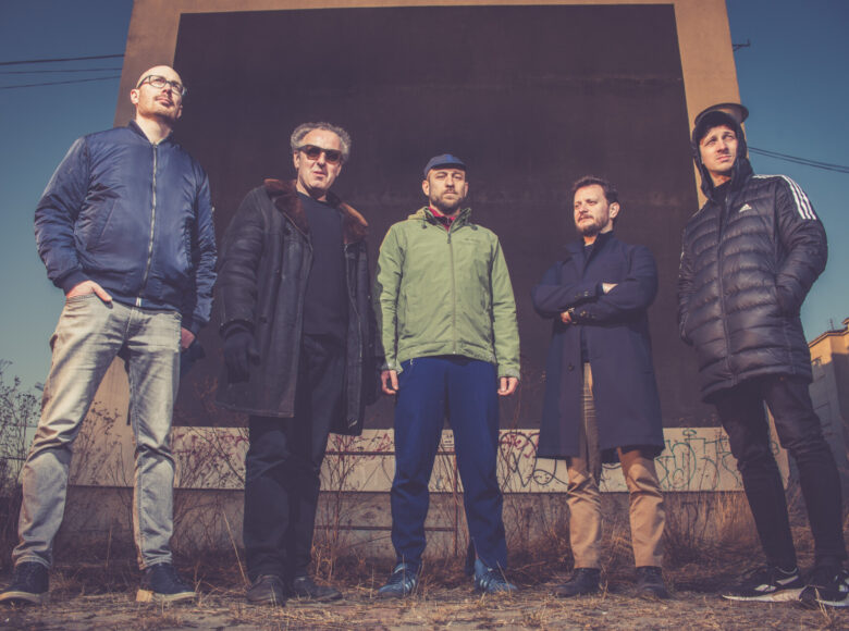 Na barwnym zdjęciu znajduje pięcioro stojących mężczyzn. Patrzą przed siebie. W tle jest blok z wielkim czarnym kwadratem.