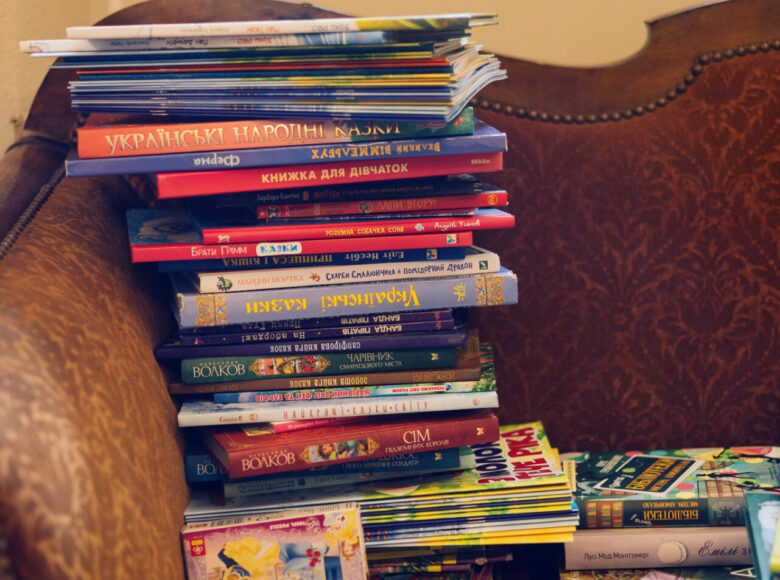 Zdjęcie. Na pomarańczowej kanapie leży stos książek. maja kolorowe okładki, tytuły książek są zapisane cyrylicą. Książki leżą również na parapecie.