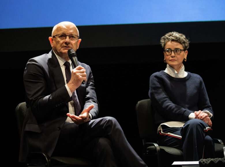 Mezczyzna w garniturze i okualarach siedzi na krześle. Mówi do mikrofonu. obok niego siedzi kobieta.