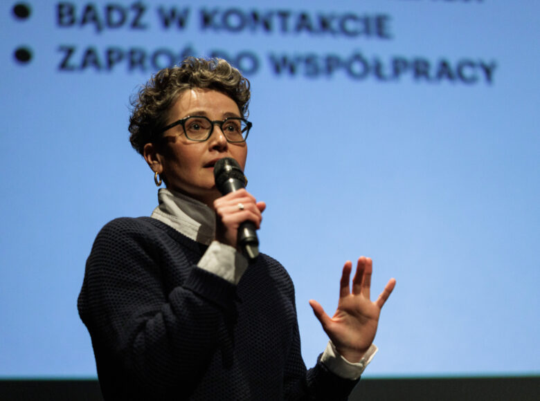 Zdjęcie. Kobieta w krótkich kręconych włosach i okularach stoi na tle białego ekranu. Mówi do mikrofonu.