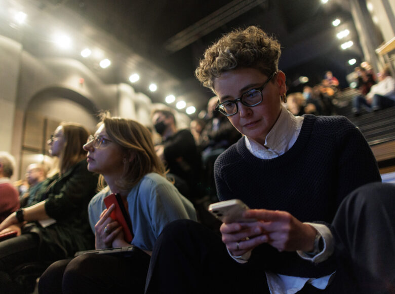 Zdjęcie. Kobieta w okularach siedzi na widowni. W rękach trzyma telefon.