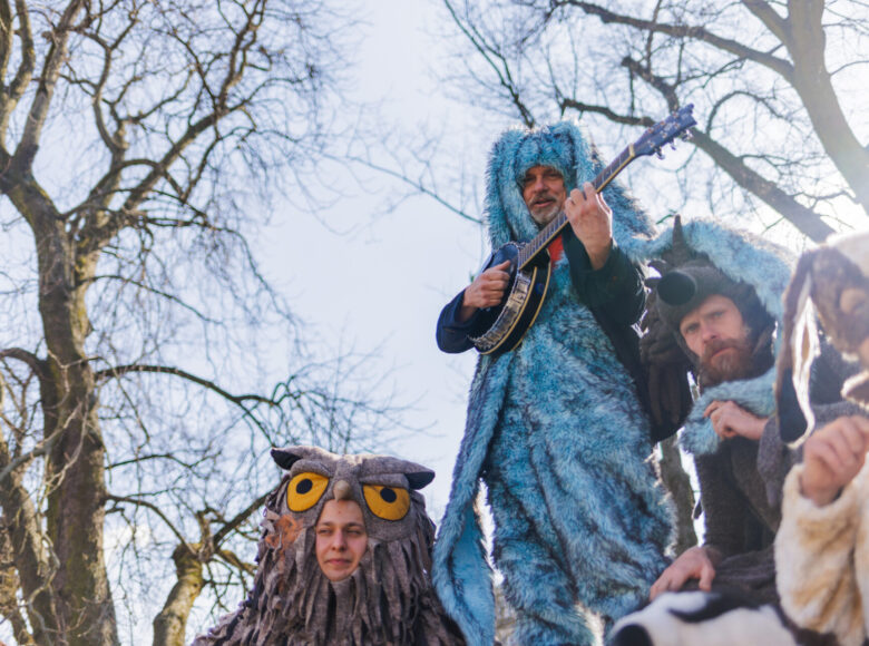 Zdjęcie. Grupa aktorów w kostiumach znajduje się z miejskim parku. Są przebrani za sowę, zająca, jeża i psa. Zając stoi wyżej od pozostałych, gra na banjo.
