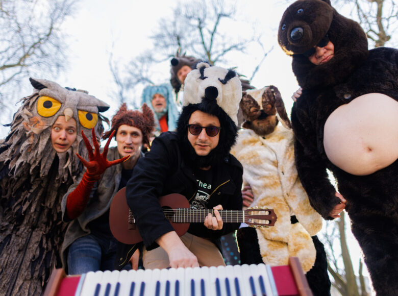 Zdjęcie. Grupa aktorów w kostiumach znajduje się z miejskim parku. Są przebrani za zwierzęta: sowę, ptaka, zająca, borsuka, jeża, psa i niedźwiedzia. Aktor przebrany za borsuka trzyma ukulele.