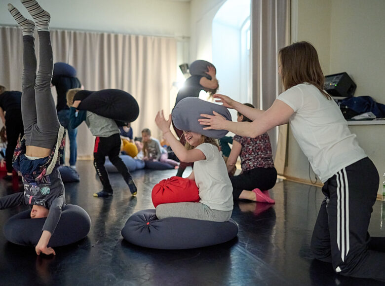 Kolorowa fotografia. Na podłodze, wśród kolorowych poduszek bawią się dzieci i dorośli. Z przodu dziewczynką stoi na głowie w poduszce, rękami podpiera się o podłogę, ma wyprostowane nogi do góry. Obok niej mama rzuca w swoją córkę miękką poduszką.