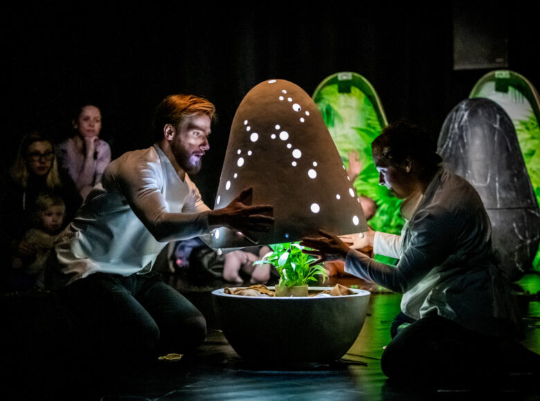 Panuje półmrok. Aktor i aktorka unoszą górną część przedmiotu przypominającego jajko. Jego środek jest oświetlony, znajduje się w nim zielona roślina.