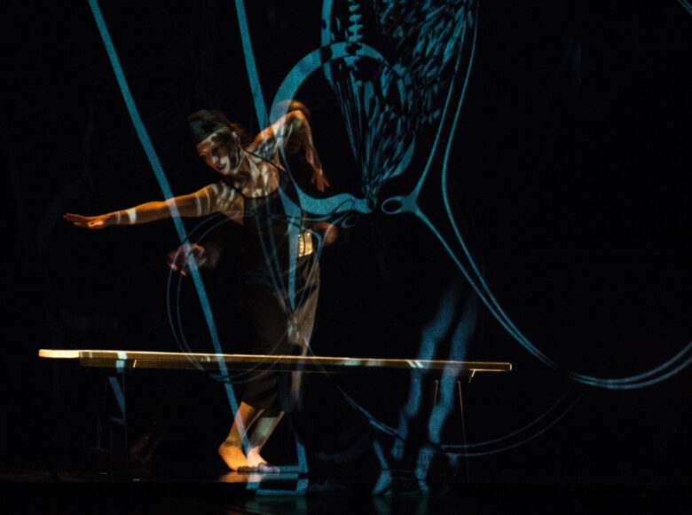 Kolorowa fotografia. Ciemna sceneria z rozproszonym światłem. Na scenie kobieta ubrana na czarno. Wygląda jakby była w trakcie wykonywania ruchów tanecznych. Przed nią stoi długa ławka bez oparcia.