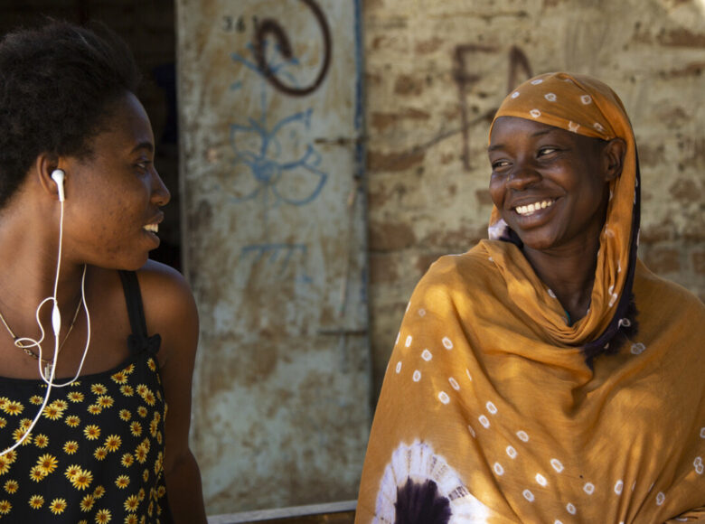 Dwie młode czarnoskóre dziewczyny, siedzą obok siebie, patrząc na siebie. Jedna w uszach ma słuchawki, druga ma głowę i ramiona okryte wzorzystą, żółtą chustą.