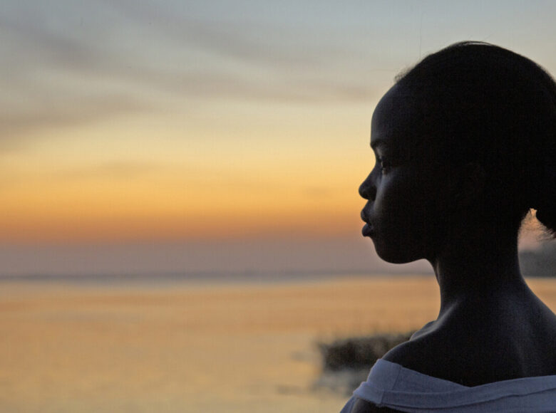 Młoda czarnoskóra dziewczyna na zbliżeniu, widziana z profilu na tle zachodzącego nad morzem słońca.