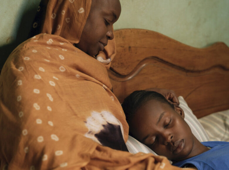 Na zdjęciu kobieta i chłopiec. Kobieta siedzi oparta o drewniany zagłówek łóżka, chłopiec leży oparty o nią, z zamkniętymi oczami.