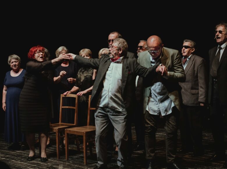 Zdjęcie. Aktorzy Teatru Seniora znajdują się na scenie. Stoją w dwóch rzędach. Na samym przodzie stoi mężczyzna, dwie osoby ciągną go za ręce.
