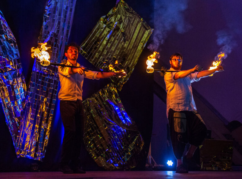 Zdjęcie. Na scenie znajduje się dwóch mężczyzn. Wykonują pokaz z użyciem płonących pochodni.