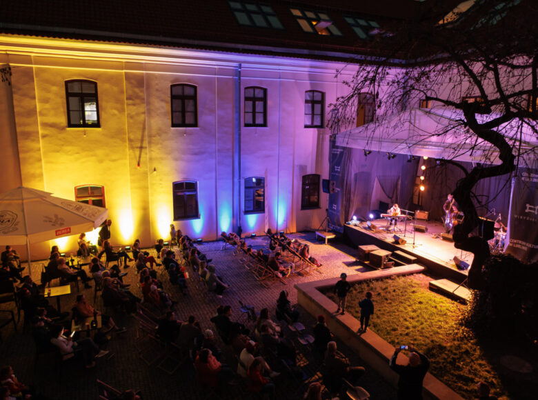 Zdjęcie Wirydarz Centrum Kultury po zmroku. Ujęcie z góry. Widzowie siedzą na krzesłach i leżakach i słuchają koncertu. Ściany wirydarza podświetlone są na niebieski i żółty.