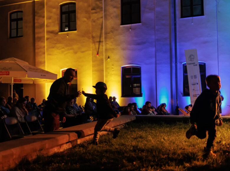 Zdjęcie. Wirydarz Centrum Kultury w Lublinie po zmroku. Ściany podświetlone są w kolorach niebieskim i żółtym. Przed sceną biegają małe dzieci.