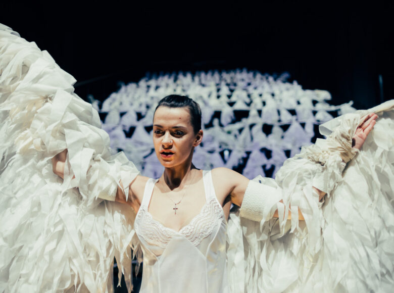 Zdjęcie. Scena ze spektaklu "Dziady". Kobieta w stroju anioła unosi ręce imitując ruch skrzydeł. Za nią, na miejscu widowni znajdują się białe kukły.