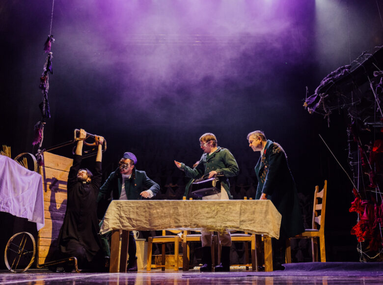 Zdjęcie. Scena ze spektaklu "Dziady". Za stołem nakrytym lnianym obrusem znajdują się mężczyźni w wojskowych płaszczach. Do drewnianej belki przywiązany za ręce jest ksiądz.